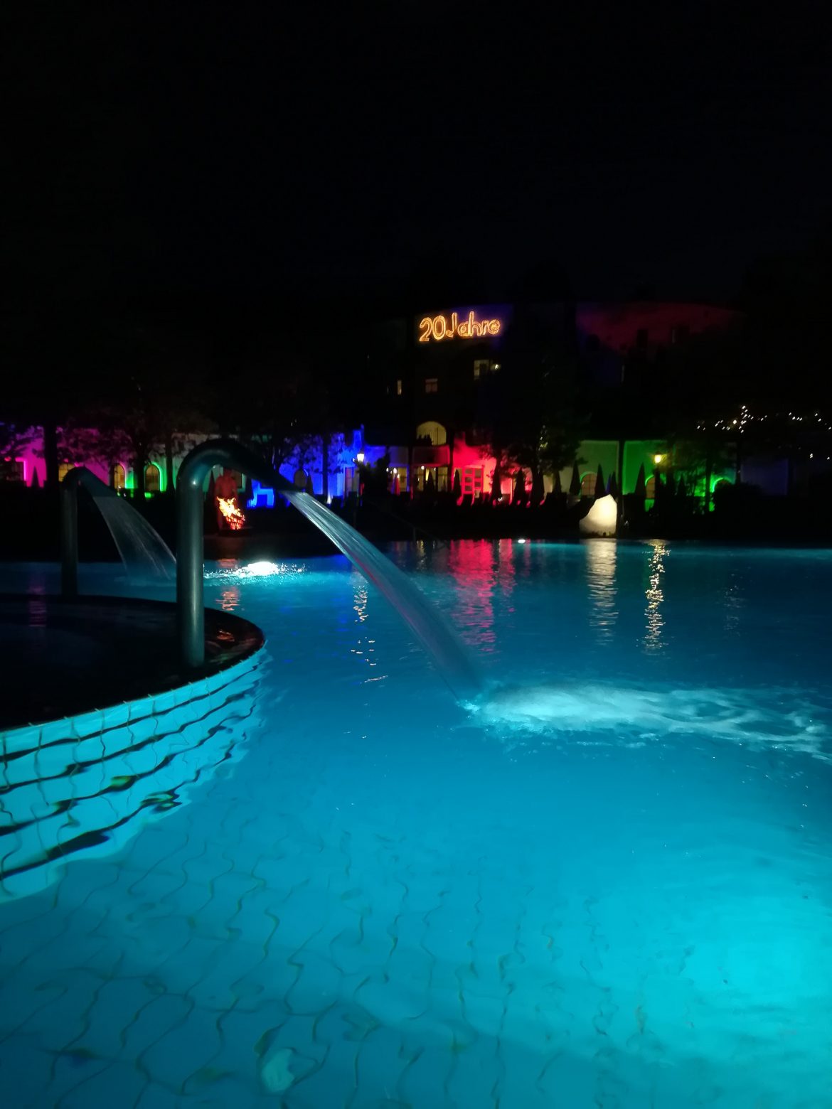Das blaue Aussenbecken der Therme Bad Blumau, mit der bunten Beleuchtung und dem Schriftzug, 20 Jahre, im Hintergrund