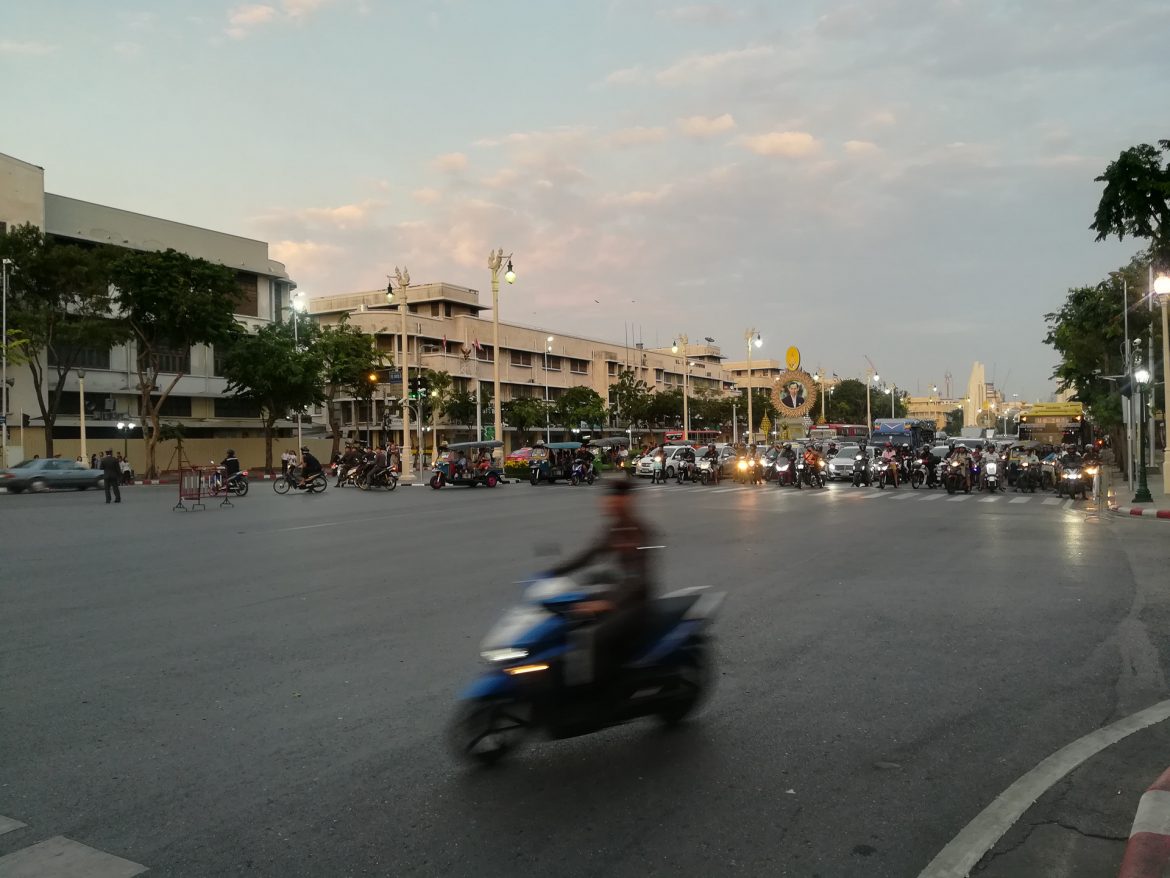 Ein einzelnes Moped fährt mitten durch das Bild, während eine ganze Horde an der roten Ampel der Kreuzung wartet.