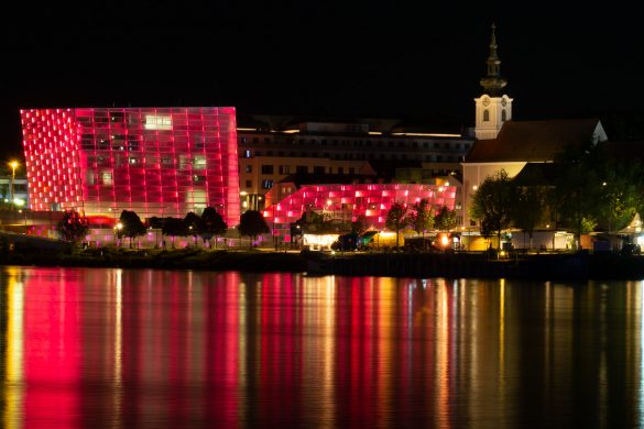 Das Ars Electronica Center hell erleuchtet in roten Farben, die sich auf der Donau spiegeln.