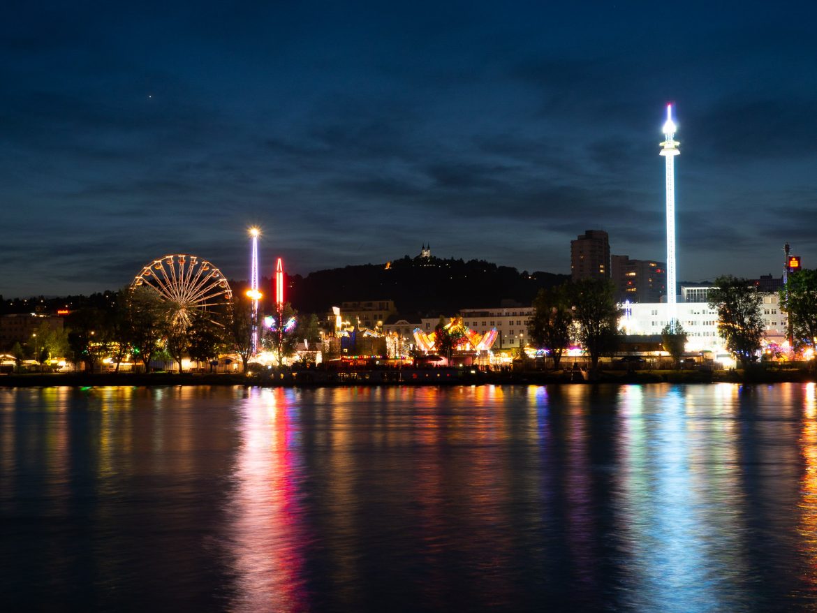 Der Urfahraner Markt in Linz, fotografiert vom gegenüberliegendem Flussufer mit den Reflektionen der Lichter im Wasser.