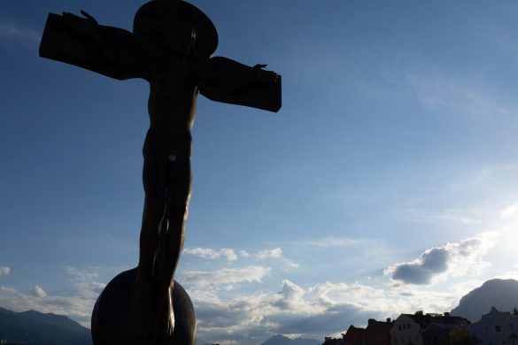 Eine Statue von Jesus am Kreuz im Sonnenschein, mit den Häusern von Innsbruck und einem leicht bewölkten Himmel im Hintergrund.