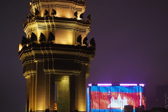 Das Independence Monument in Phnom Penh, aufgenommen in der Nacht, wärend im Hintergrund eine große Reklame die Flagge von Kambodscha zeigt.