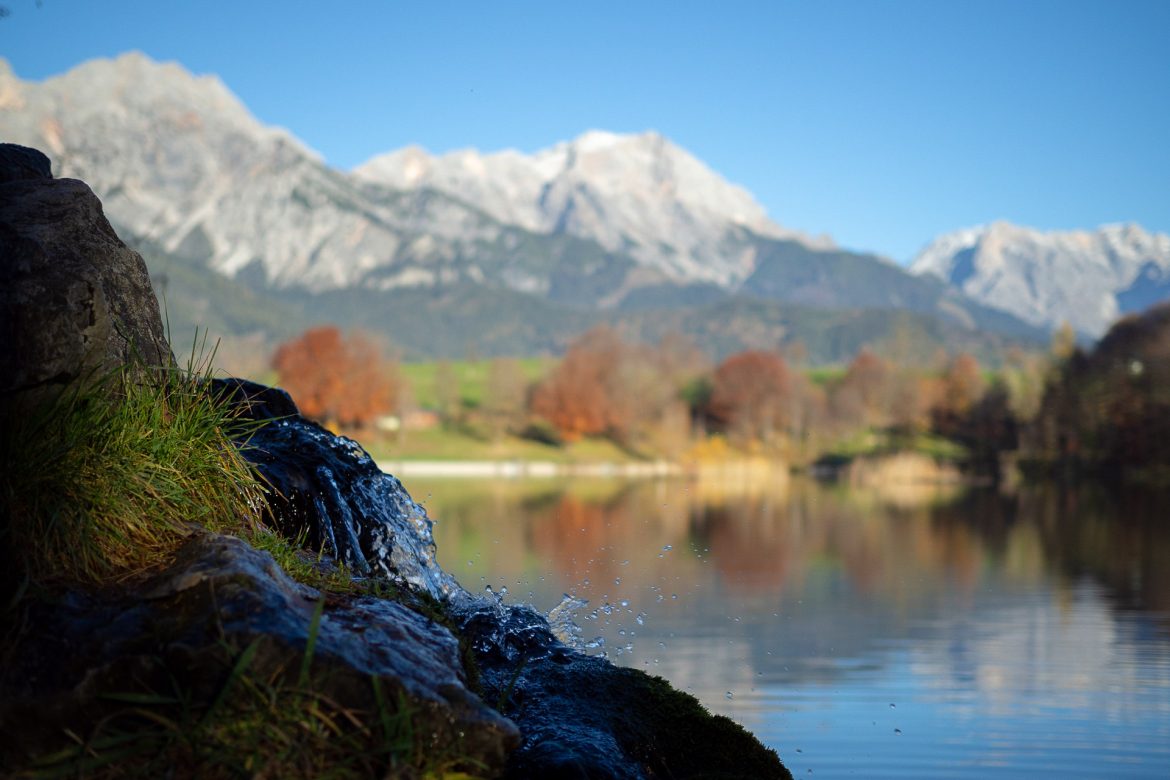 Ein kleiner Zufluss zum Ritzensee plätschert leise auf einen Stein während sich im Hintergrund die die Herbstbäume im See spiegeln und die Berge über sie hinausragen.