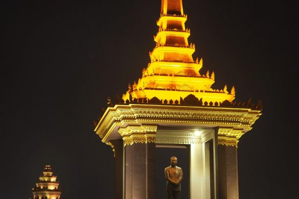Eine Statue von Samdech Chuon Nath in einer Stupa und dem National Memorial im Hintergrund, beleuchtet bei Nacht