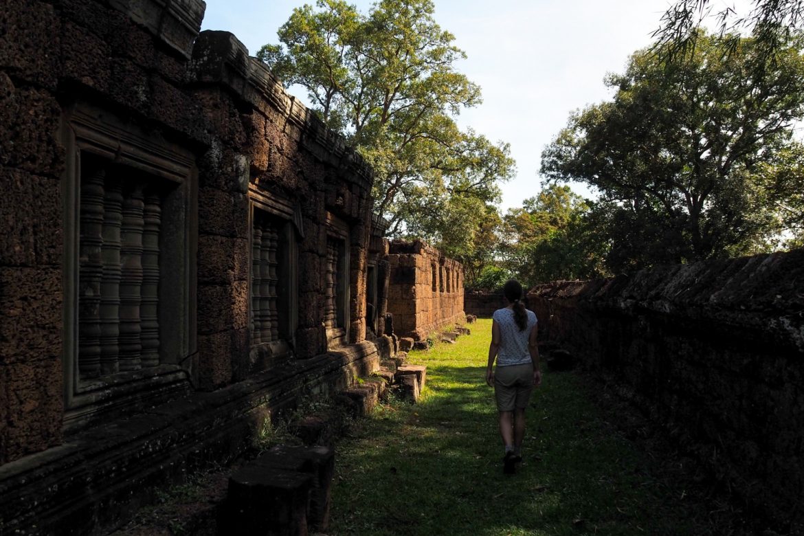 Anita fotografiert von hinten, während sie durch den äußeren Bereich von East Mobon geht. Links und rechts sieht man die alten Mauern des Tempels mit den Fenstern und Säulen darin.