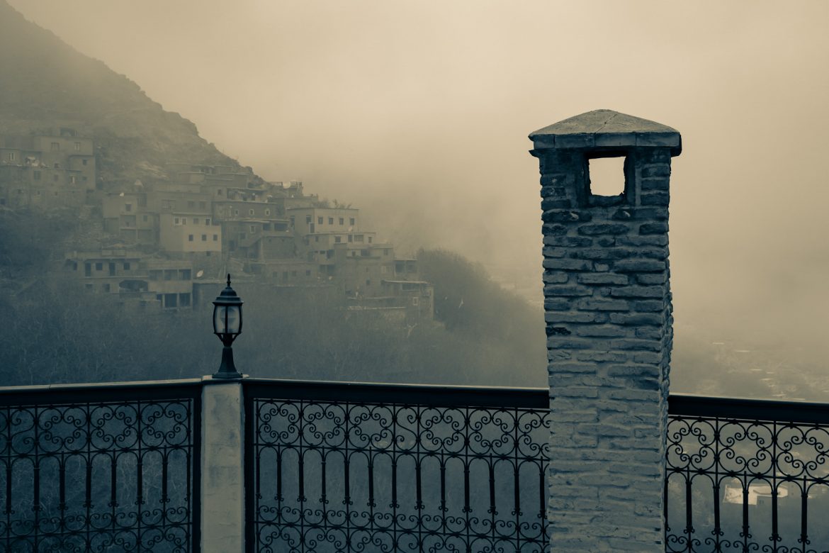 Eine Lampe und ein kleiner Turm auf dem Balkon der Kasbah von Imlil im Vordergrund während sich der Nebel über ein paar Häuser zieht, die in den Berghang gebaut wurden. Foto in schwarz-weiß.