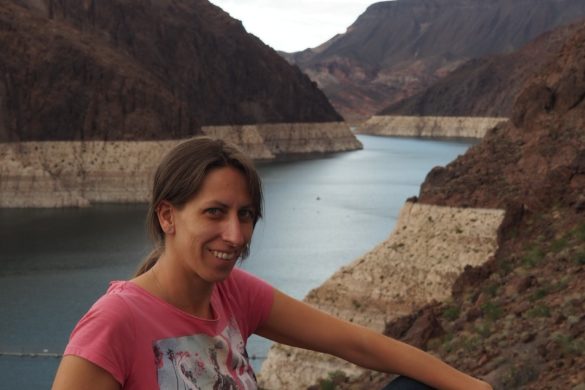 Anita sitzt auf einer der Mauern rund um den Hoover Dam mit dem Stausee im Hintergrund und den Felsen, an denen man den üblichen weit höheren Wasserstand ablesen kann.