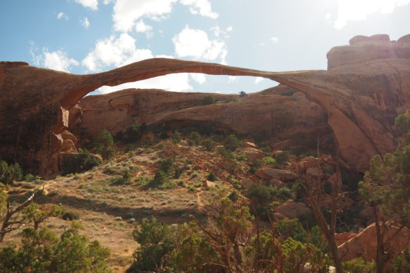 Der Landscape Arch erhebt sich über die Landschaft im Arches National Park mit einem leicht bewölkten Himmel im Hintergrund und einigen Büschen und kleinen Bäumen im Vordergrund